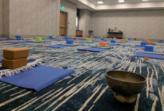 Carpeted hotel conference room set up for group yoga and meditation with rows of blue mats, colorful foam blocks and blankets, and a singing bowl in the foreground.