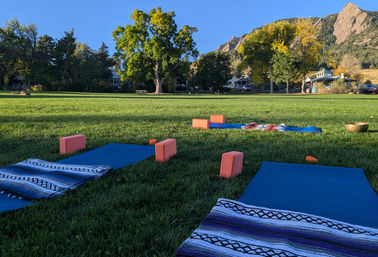 Blue yoga mats with striped blankets and coral yoga blocks arranged on a grassy park lawn, ready for sunrise yoga, framed by large trees and rocky mountain foothills under a clear blue sky.