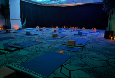 Calm, candlelit indoor yoga setup in a dim event room with rows of blue mats, yoga blocks, blankets and warm LED candles for meditation