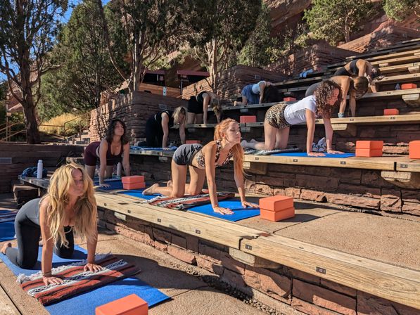Outdoor group yoga class on terraced wooden benches at a red sandstone amphitheater, people on blue mats and orange blocks practicing cat-cow stretches under sunny skies and trees.
