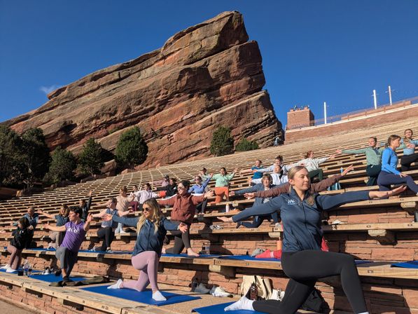 Sunlit group yoga on blue mats at a red-sandstone outdoor amphitheater, participants stretching on stepped stone benches beneath a clear blue sky.