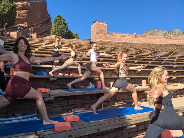 Smiling women doing outdoor yoga warrior poses on blue mats in a sunlit red-rock stone amphitheater with tiered wooden benches and a clear blue sky.