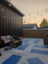 Cozy suburban backyard patio at sunset with hanging string lights, blue yoga mats laid on concrete, outdoor sofa, patterned rug and a glowing fire pit.
