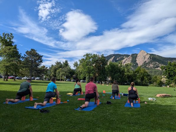 Sunlit outdoor yoga class on blue mats in a green park, participants stretching toward dramatic rocky foothills and a bright blue sky with wispy clouds.