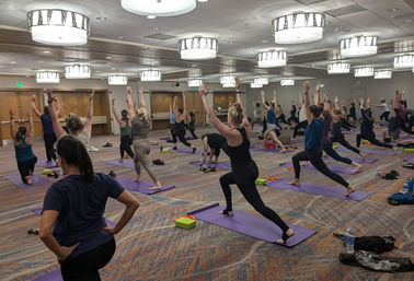 Group yoga class in a hotel conference room with many participants on purple mats practicing Warrior I lunges under round ceiling lights — indoor fitness workshop