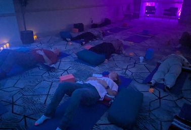 Candlelit restorative yoga and guided meditation session in a dim purple-blue studio, participants lying on mats with bolsters, blocks and soft lighting.