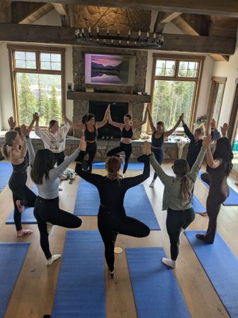 Group yoga class in a rustic mountain lodge, participants in tree pose on blue mats around a stone fireplace with large windows overlooking a pine forest.