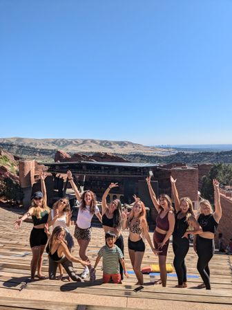 Happy group of women and a child posing with arms raised on wooden tiers of a red‑rock outdoor amphitheater in Colorado, stage and rolling hills with distant city skyline under a clear blue sky