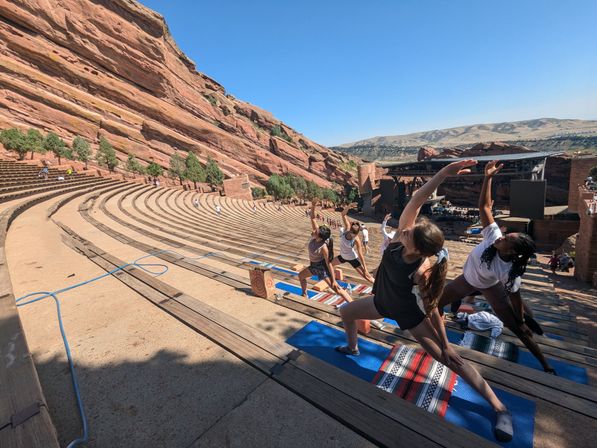 Group doing yoga on mats across tiered wooden benches in a sunlit red‑rock outdoor amphitheater, dramatic canyon walls and stage under a clear blue sky