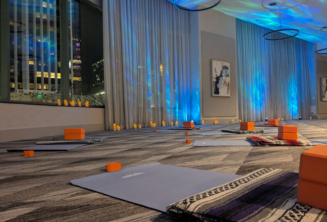 Evening wellness setup in a city conference room with yoga mats, orange blocks, patterned blankets and faux candles on a carpeted floor, blue uplighting and floor-to-ceiling windows showing city buildings