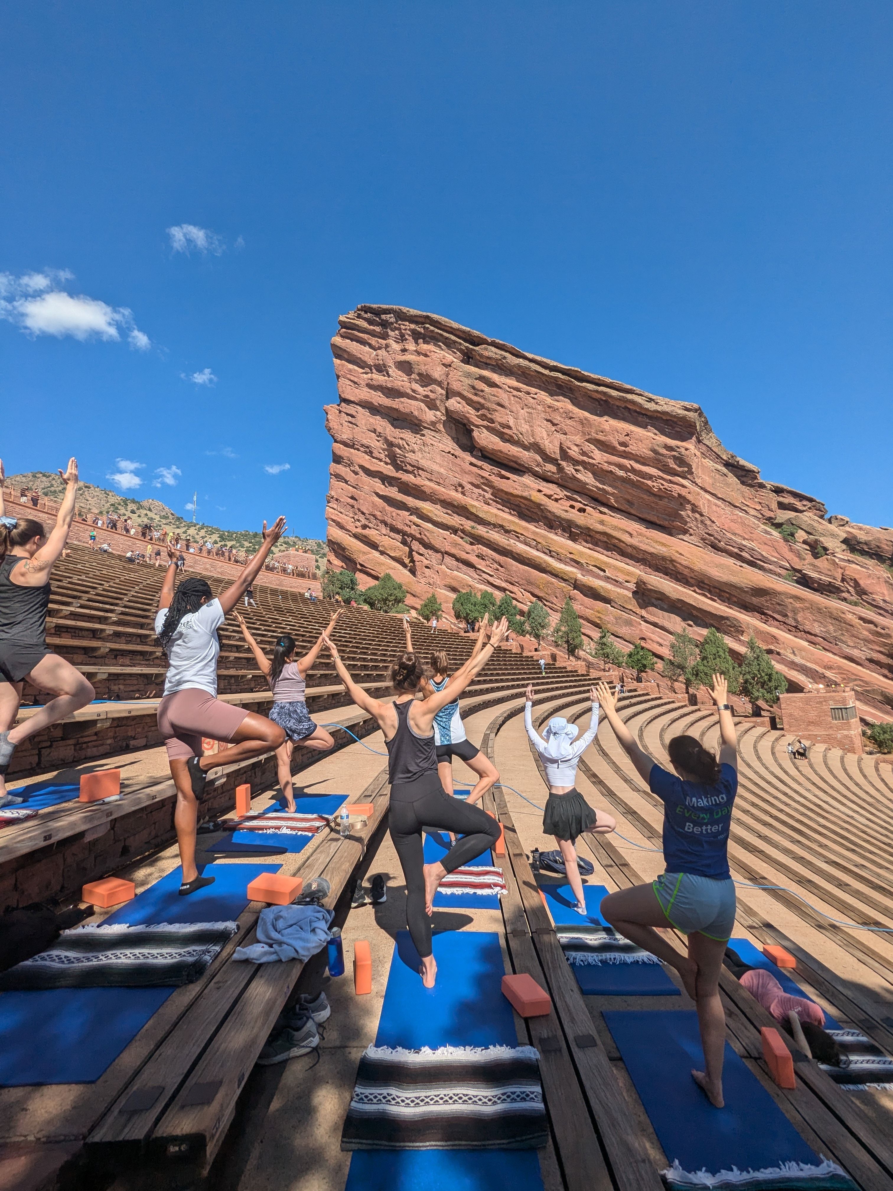 Group outdoor yoga on blue mats at a red sandstone amphitheater in Colorado, students holding tree pose against a dramatic rock formation and clear blue sky.