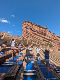 Group outdoor yoga on blue mats at a red sandstone amphitheater in Colorado, students holding tree pose against a dramatic rock formation and clear blue sky.