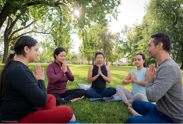 Five adults seated in a circle on yoga mats in a sunny green park, eyes closed with hands in prayer pose during a peaceful outdoor meditation session beneath leafy trees.
