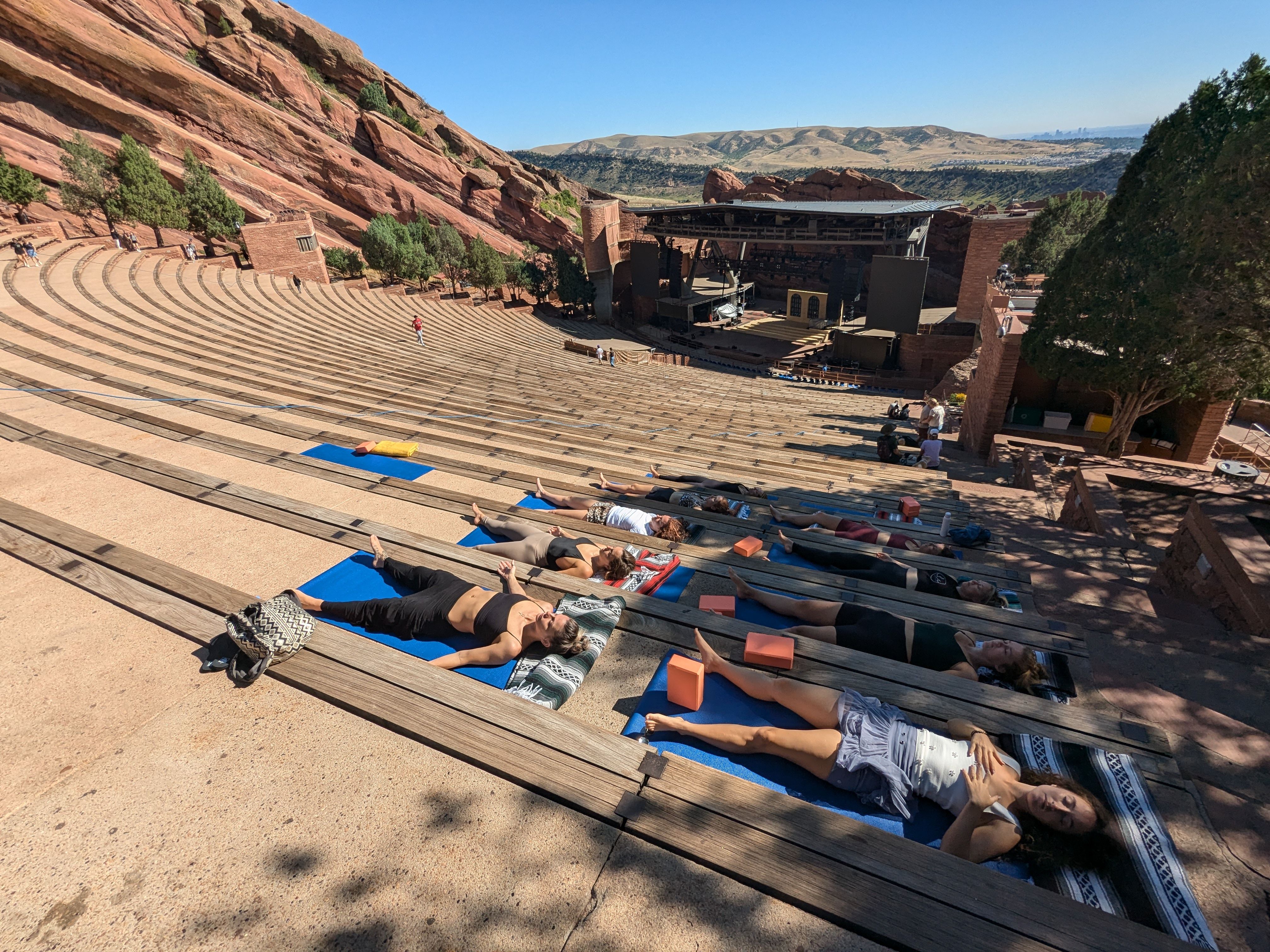 Outdoor yoga session with people lying on mats across terraced wooden benches of a red‑rock amphitheater, surrounded by rocky cliffs and wide mountain views under a clear blue sky.