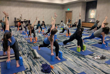 Indoor group yoga class on blue mats in a carpeted room, participants using yoga blocks and blankets while holding a twisting lunge with arms raised — community fitness session.