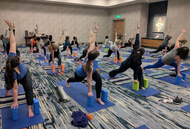 Indoor group yoga class on blue mats in a carpeted room, participants using yoga blocks and blankets while holding a twisting lunge with arms raised — community fitness session.
