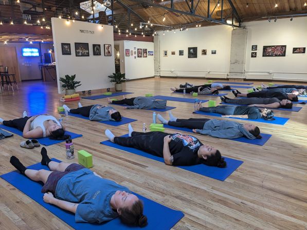 Group yoga class in Savasana on blue mats in a spacious loft-style studio with hardwood floors, exposed beams, string lights and artwork on the walls.