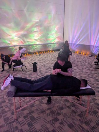 Person receiving a massage on a table in a dim wellness studio with aurora-style green and purple light projections on the walls, flickering LED candles along the perimeter and others getting chair massages in the background.