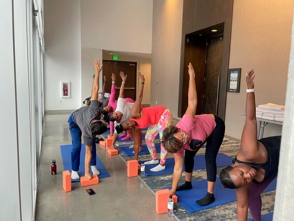 Group yoga class of women performing a standing side-bend/triangle pose on blue mats with orange blocks and water bottles in a bright indoor studio