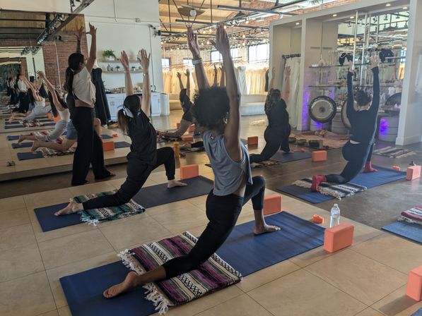 Group yoga class practicing low lunge in a bright urban studio with exposed brick, floor-to-ceiling mirrors, mats, blocks, blankets, and a gong.