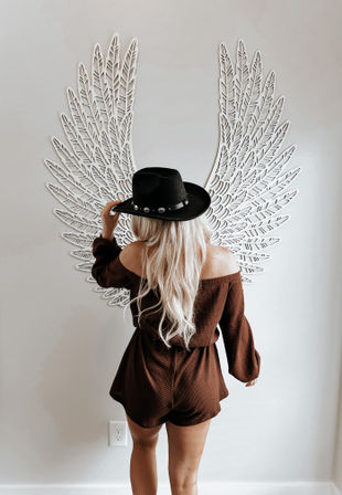 Blonde woman with back to camera wearing a black hat and brown off-shoulder romper, posing like an angel in front of large white decorative wings wall art in a neutral indoor setting