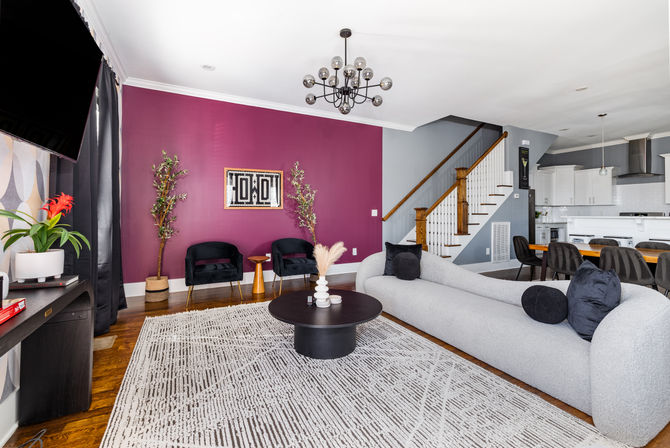 Modern open-plan living room with a plum accent wall, curved light-gray sofa, black round coffee table on a patterned rug, two velvet armchairs, mid-century chandelier, wooden staircase, and a white kitchen/dining area in the background.