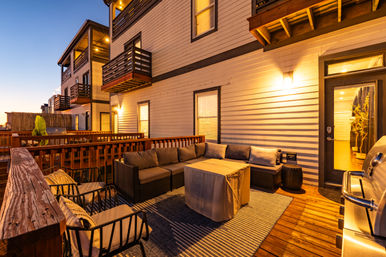 Cozy twilight townhouse deck patio with wicker sectional sofa, cushioned chairs, covered coffee table, gas grill and wooden railings lit by warm exterior lights.