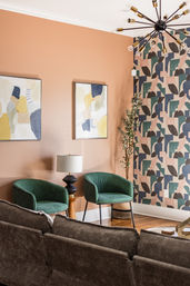 Cozy modern living room corner with terracotta accent wall, geometric wallpaper, two green upholstered chairs, wooden side table with black lamp, abstract art and mid-century sputnik chandelier.