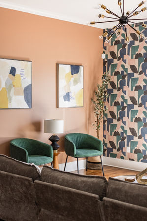 Cozy modern living room corner with terracotta accent wall, geometric wallpaper, two green upholstered chairs, wooden side table with black lamp, abstract art and mid-century sputnik chandelier.