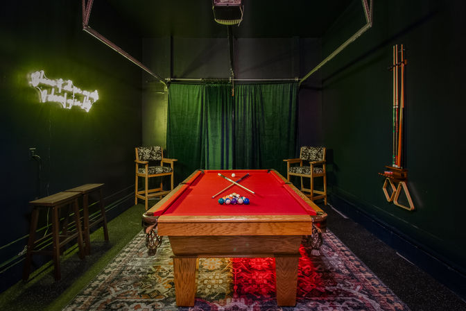 Red-felt pool table centered in a cozy indoor game room with neon wall sign, green curtains, patterned rug, two wooden armchairs, bar stools and wall-mounted cue rack.