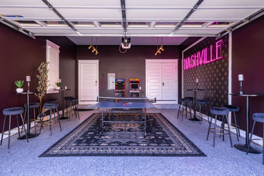 Converted garage game room in Nashville featuring a ping-pong table on a patterned rug, two retro arcade cabinets, velvet bar stools and a pink neon "NASHVILLE" sign