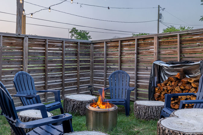 Cozy backyard fire pit at dusk with a metal fire bowl and flickering flames, blue Adirondack chairs and tree-stump seats, string lights overhead and stacked firewood against a wooden privacy fence.