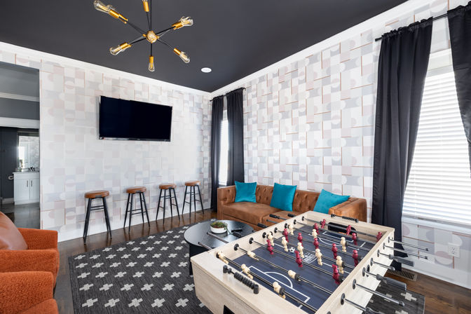 Playful modern game room with foosball table in the foreground, leather sectional with teal pillows, wall-mounted TV above a row of wooden bar stools, geometric wallpaper, black sputnik light fixture, patterned rug and tall windows with dark curtains.