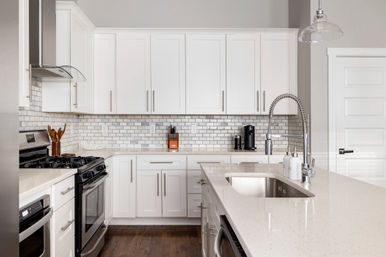 Bright modern white kitchen with quartz island, undermount stainless sink and spring pull-down faucet, white subway tile backsplash, shaker cabinets, gas range and stainless appliances on dark hardwood floors