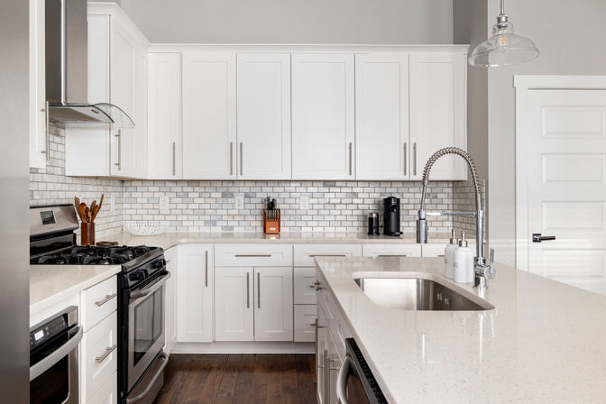 Bright modern white kitchen with quartz island, undermount stainless sink and spring pull-down faucet, white subway tile backsplash, shaker cabinets, gas range and stainless appliances on dark hardwood floors