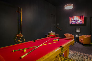 Red-felt pool table in a cozy game room with cues on a wall rack, leather lounge chairs, barrel side table, wall-mounted TV showing a crackling fireplace, and a sparkling chandelier