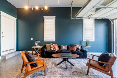 Stylish garage lounge with deep teal accent wall, black velvet sofa with patterned throw pillows, two mid-century leather armchairs, round wooden coffee table, track lighting, potted plant and open garage door