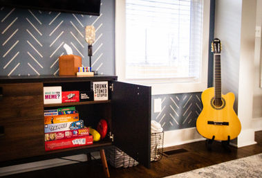 Cozy living room corner with a yellow acoustic guitar on a stand by a window with blinds, an open mid-century cabinet showing stacked board games and toys against a patterned gray accent wall and hardwood floor.