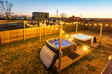 Cozy evening backyard scene: two illuminated hot tubs on a wooden deck with warm string lights in a fenced yard overlooking a distant city skyline at dusk.