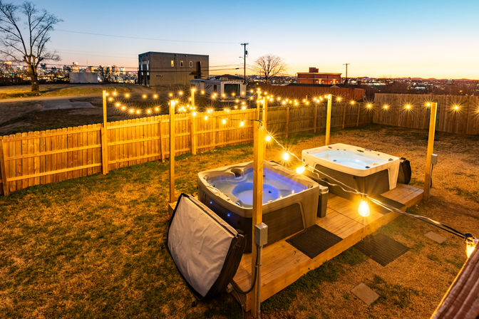 Cozy evening backyard scene: two illuminated hot tubs on a wooden deck with warm string lights in a fenced yard overlooking a distant city skyline at dusk.
