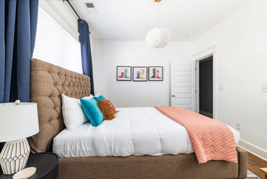 Bright modern bedroom interior with tufted brown headboard, white bedding, teal and rust accent pillows, coral quilted throw, navy curtains, bedside lamp and a white pendant light, with three framed colorful boot prints on the wall — cozy contemporary bedroom decor.