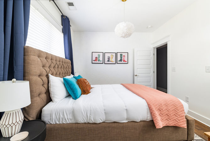 Bright modern bedroom interior with tufted brown headboard, white bedding, teal and rust accent pillows, coral quilted throw, navy curtains, bedside lamp and a white pendant light, with three framed colorful boot prints on the wall — cozy contemporary bedroom decor.