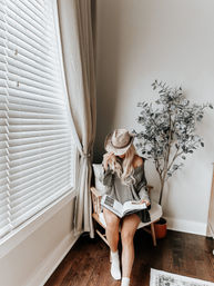 Cozy sunlit reading nook — person in an oversized sweater and straw hat reads a book on a wooden chair by white blinds, neutral curtains, potted eucalyptus, and dark hardwood floors.