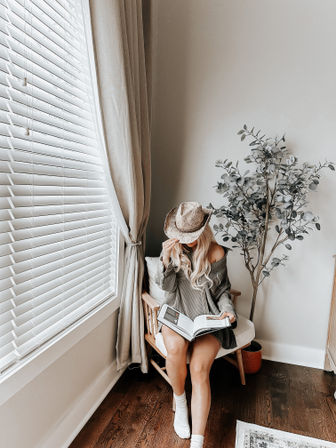 Cozy sunlit reading nook — person in an oversized sweater and straw hat reads a book on a wooden chair by white blinds, neutral curtains, potted eucalyptus, and dark hardwood floors.