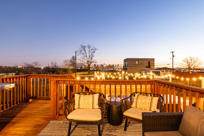 Twilight wooden deck patio with cozy cushioned chairs, patterned pillows, string lights along the railing and a distant city skyline