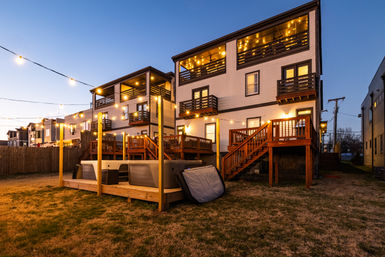 Twilight backyard scene of modern three-story townhouses with warm string lights, illuminated balconies, wooden decks and raised hot tubs on a shared platform.