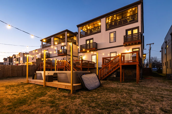 Twilight backyard scene of modern three-story townhouses with warm string lights, illuminated balconies, wooden decks and raised hot tubs on a shared platform.
