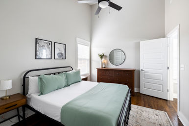 Bright modern bedroom with black metal bed frame, white sheets and sage-green pillows and throw, wooden nightstand and dresser with round mirror and vase, hardwood floors, ceiling fan, white walls and window blinds