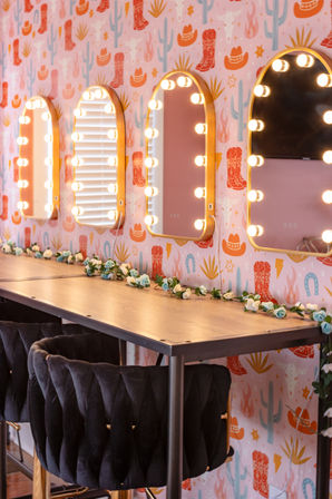 Cheerful beauty station with arched Hollywood-style vanity mirrors framed by glowing bulbs above a wooden makeup counter, floral garland and plush black velvet stools against pink cactus-and-cowboy-hat wallpaper.