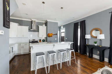 Stylish contemporary home kitchen interior with white cabinets, subway tile backsplash and stainless steel range, marble breakfast bar with four metal stools, gray walls, wood floors, pendant lights, black curtains, console table with lamps and round mirror, and a decorative guitar on the wall.